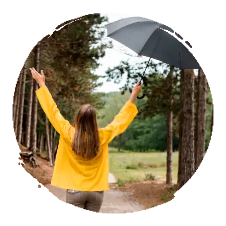  une femme habillée en jaune et gris qui marche sur un chemin dans une forêt, les bras en l'air et écartés ave cun parapluie dans une main